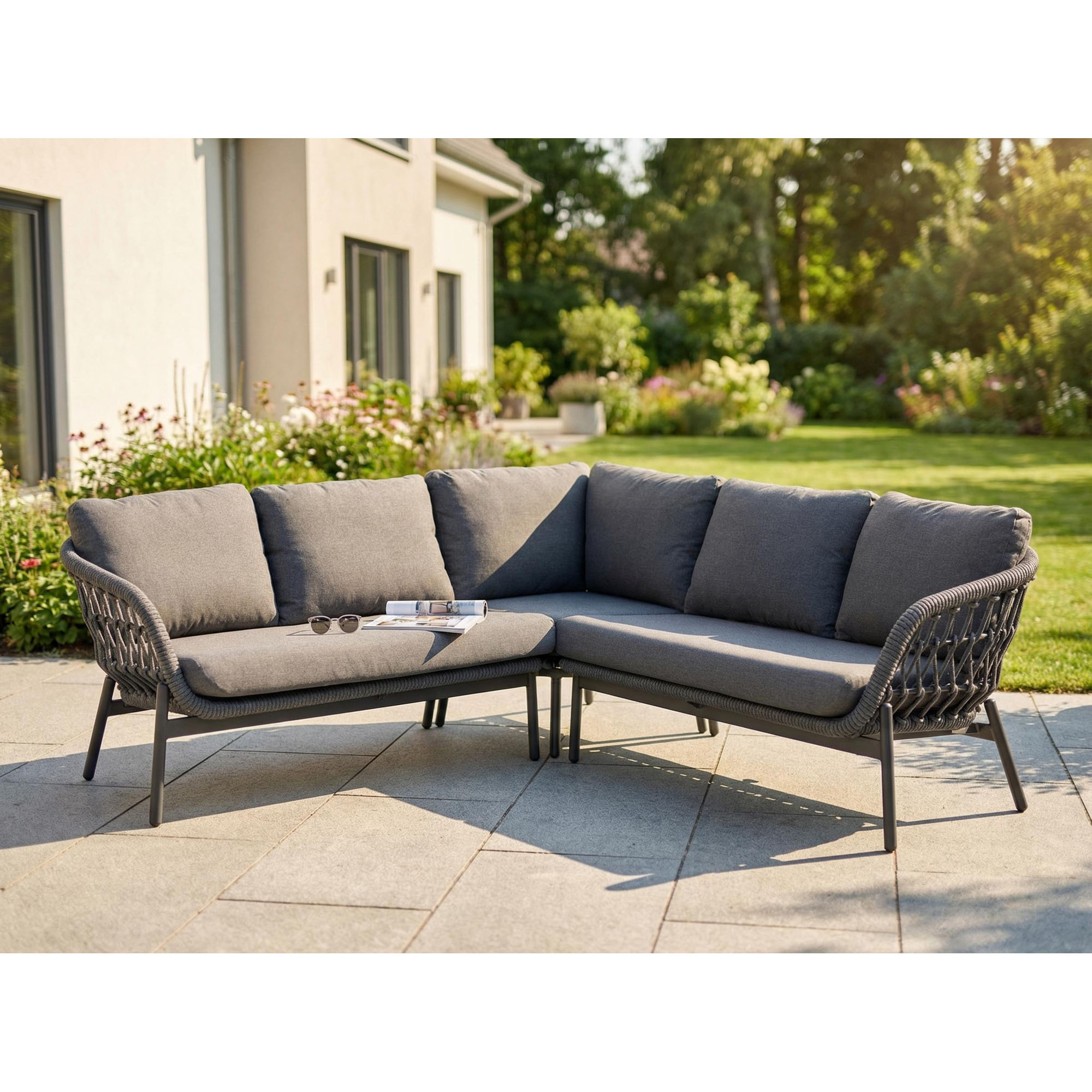 graue Gartenlounge mit Ecksofa und Kissen steht auf einer Terrasse vor einem Garten mit Bäumen und Pflanzen