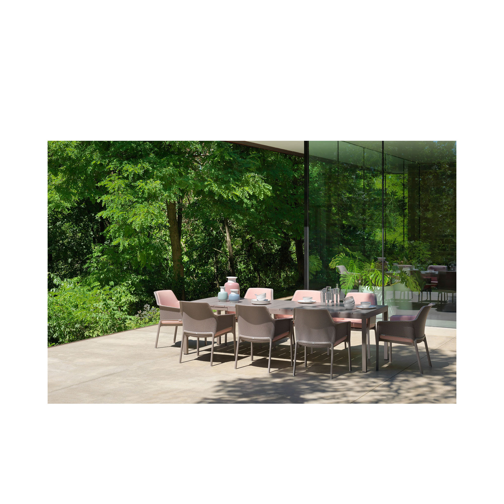 Eine moderne Terrasse mit mehreren taupefarbenen Stühlen und einem rechteckigen Esstisch. Auf dem Tisch stehen Geschirr, Gläser und Dekoration, im Hintergrund sind grüne Bäume und Glaswände zu sehen.