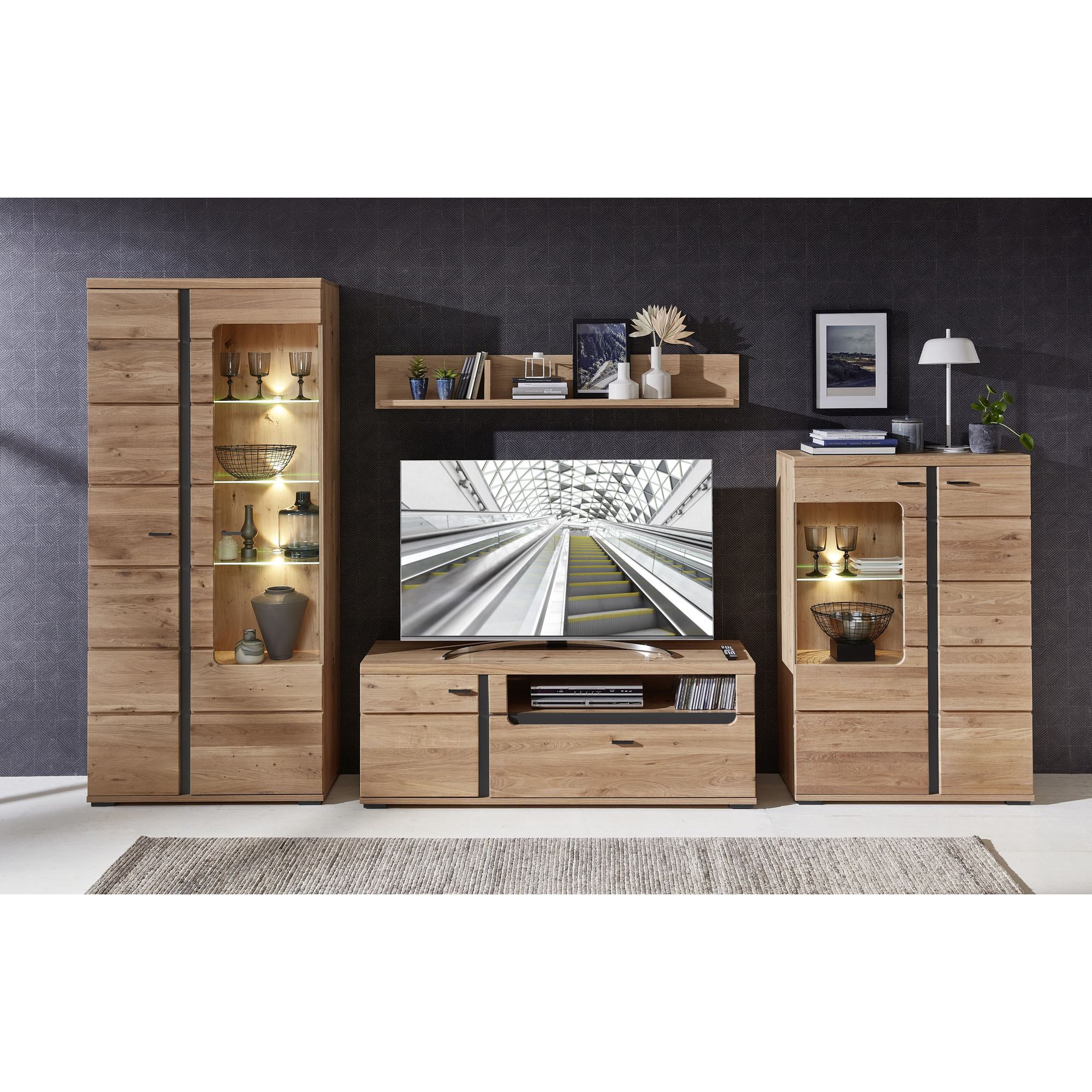 Wohnzimmeransicht mit einer Wohnwand in Holzoptik. Links und rechts stehen Vitrinen mit beleuchteten Glasfächern. In der Mitte befindet sich ein TV-Lowboard mit Fernseher. Darüber hängen zwei Wandboards mit Büchern, Vasen und Bildern.