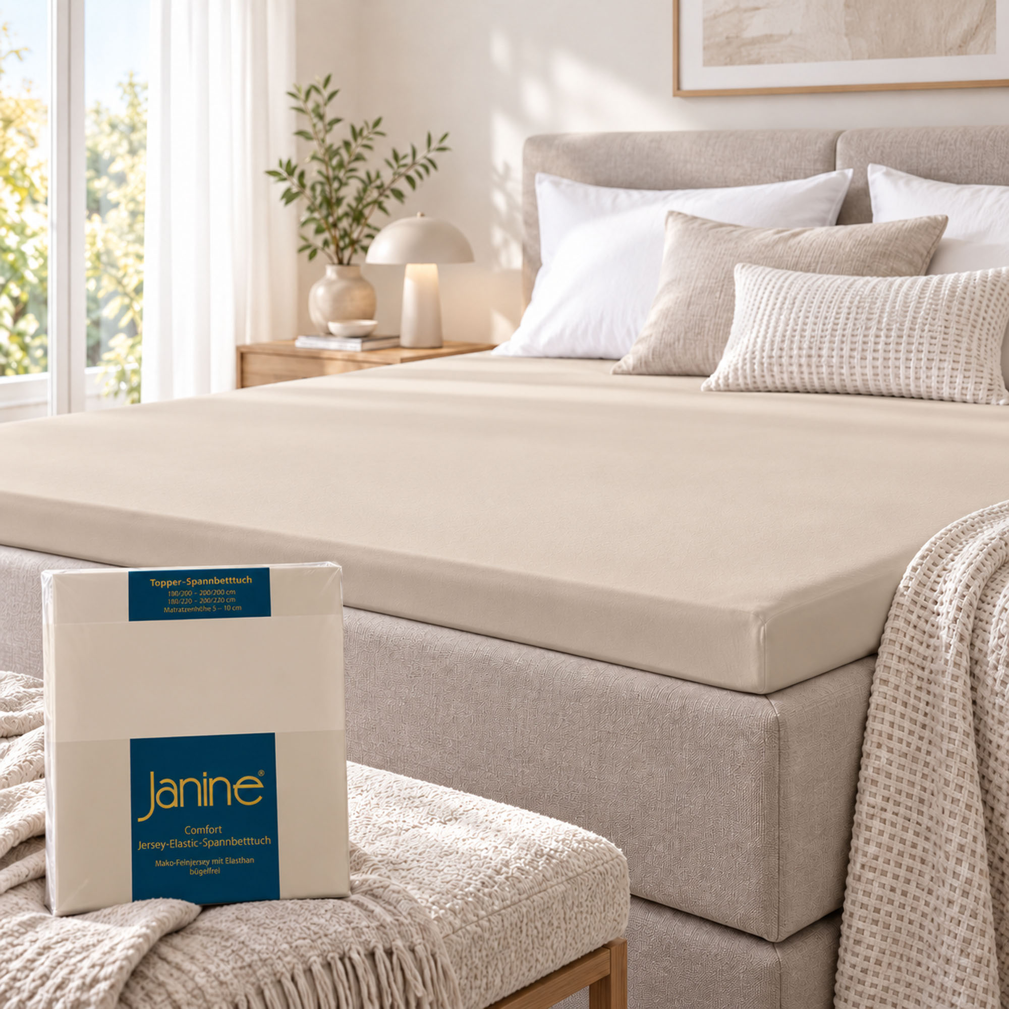Sandfarbenes Janine Topper-Spannbetttuch 5001 liegt glatt auf dem Topper eines Boxspringbetts; die Verpackung ist im Vordergrund zu sehen.