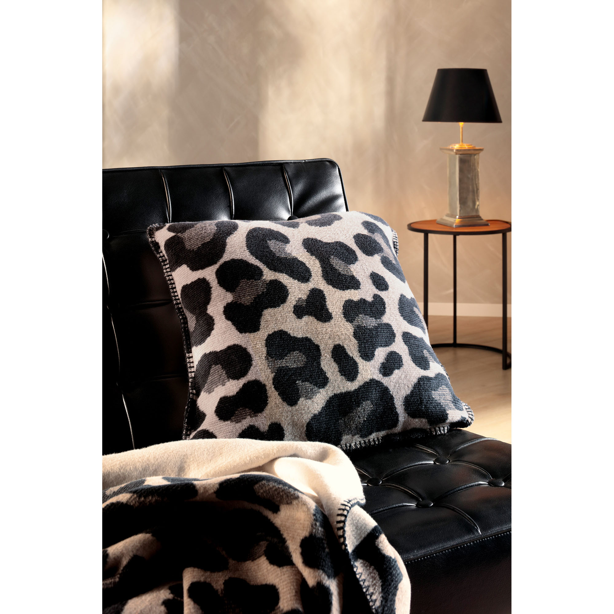 Kissenhülle Leo Part liegt auf einem schwarzen Ledersessel, davor ist eine passende Decke mit Leopardenmuster drapiert.