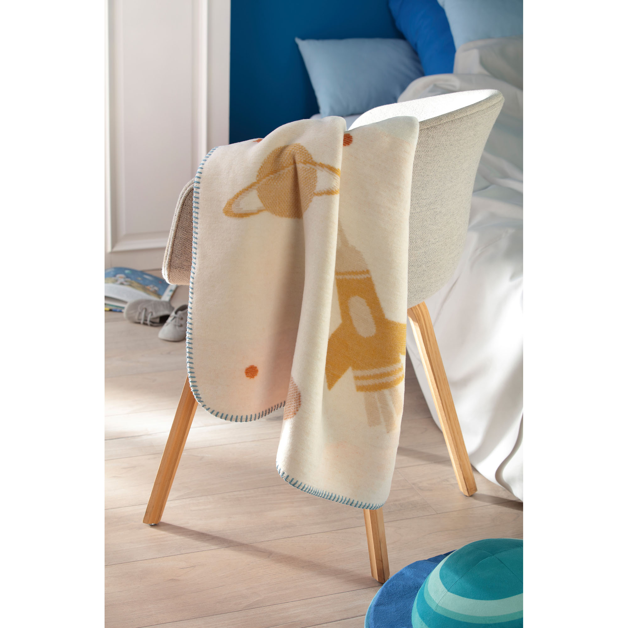 Babydecke Orbit hängt über einem Kinderstuhl im hellen Kinderzimmer und zeigt orange-blaue Weltraummotive.