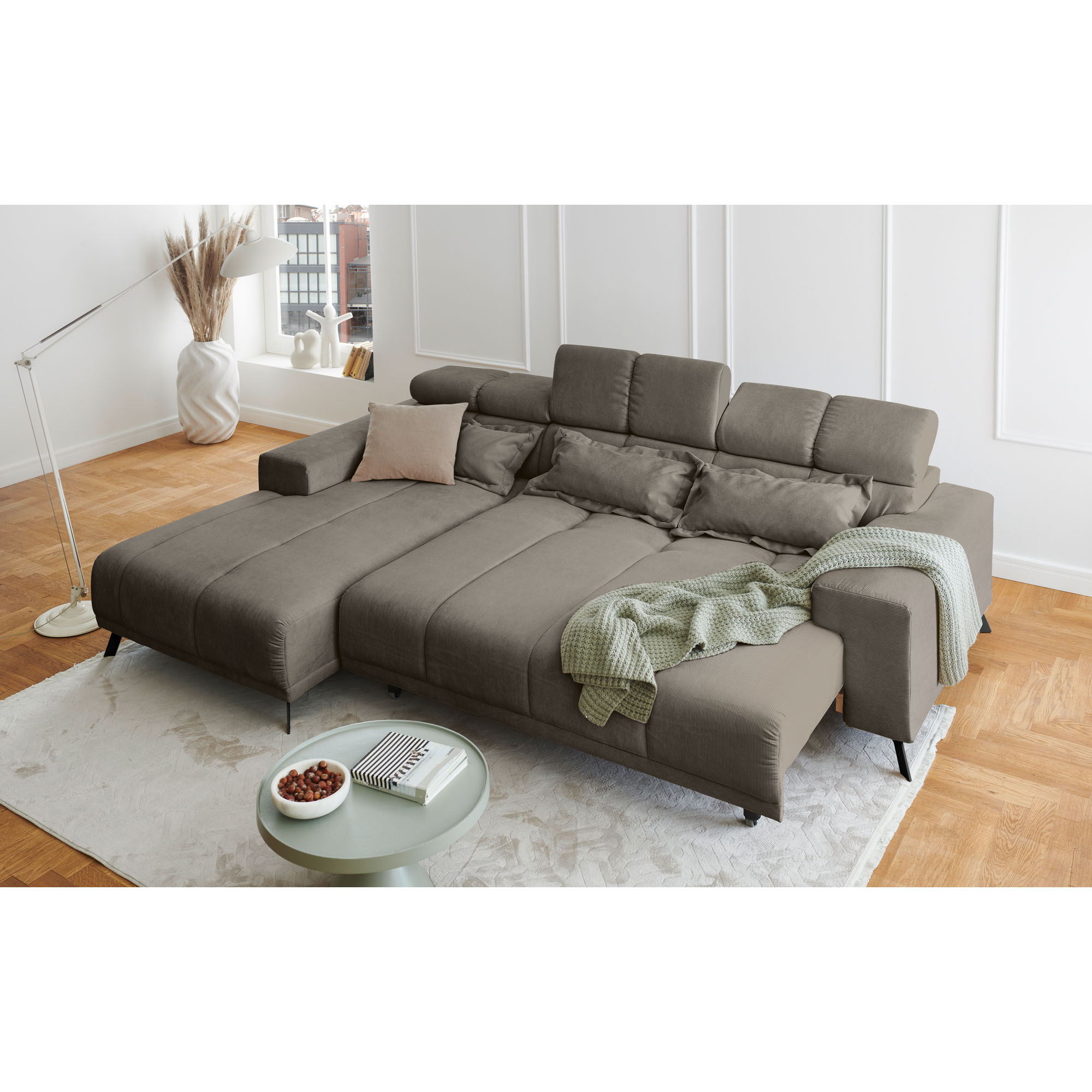 Hellgraues Ecksofa mit Ottomane links und ausgezogener Liegefläche in einem hellen Wohnzimmer mit Teppich und Couchtisch