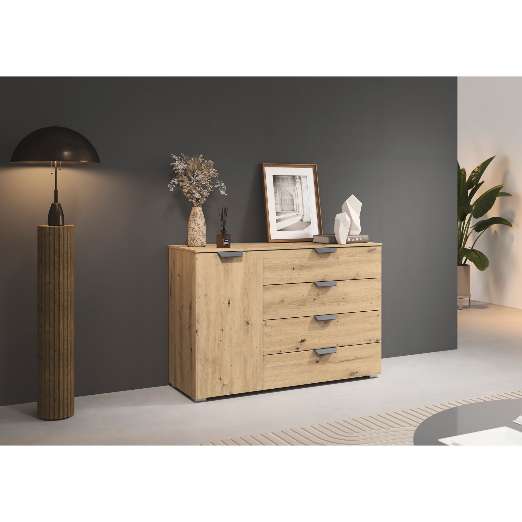 Kommode in Eiche Artisan mit vier Schubladen und einer Tür, dekoriert mit Vase, Bildrahmen und Büchern, vor dunkler Wand mit Stehlampe und Pflanze