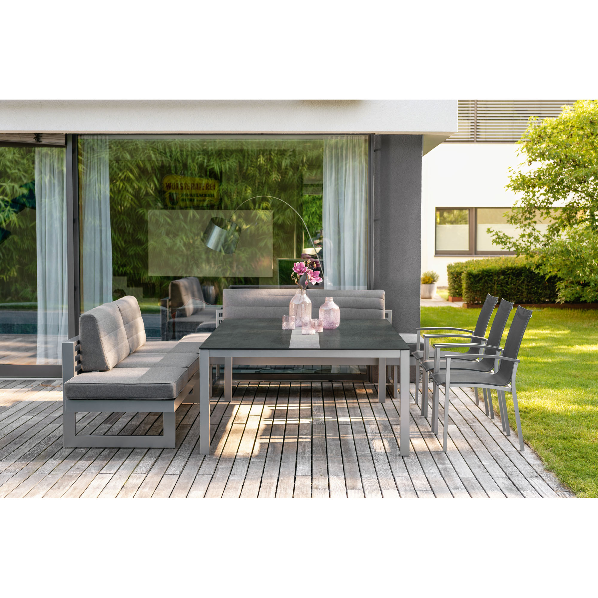 Eine Terrasse mit grauer Sitzgruppe bestehend aus zwei Bänken mit Rückenlehne, einem Tisch und mehreren Stühlen. Auf dem Tisch stehen Glasvasen mit Blumen, im Hintergrund sind Glasfronten und ein Garten mit Rasen und Bäumen zu sehen.