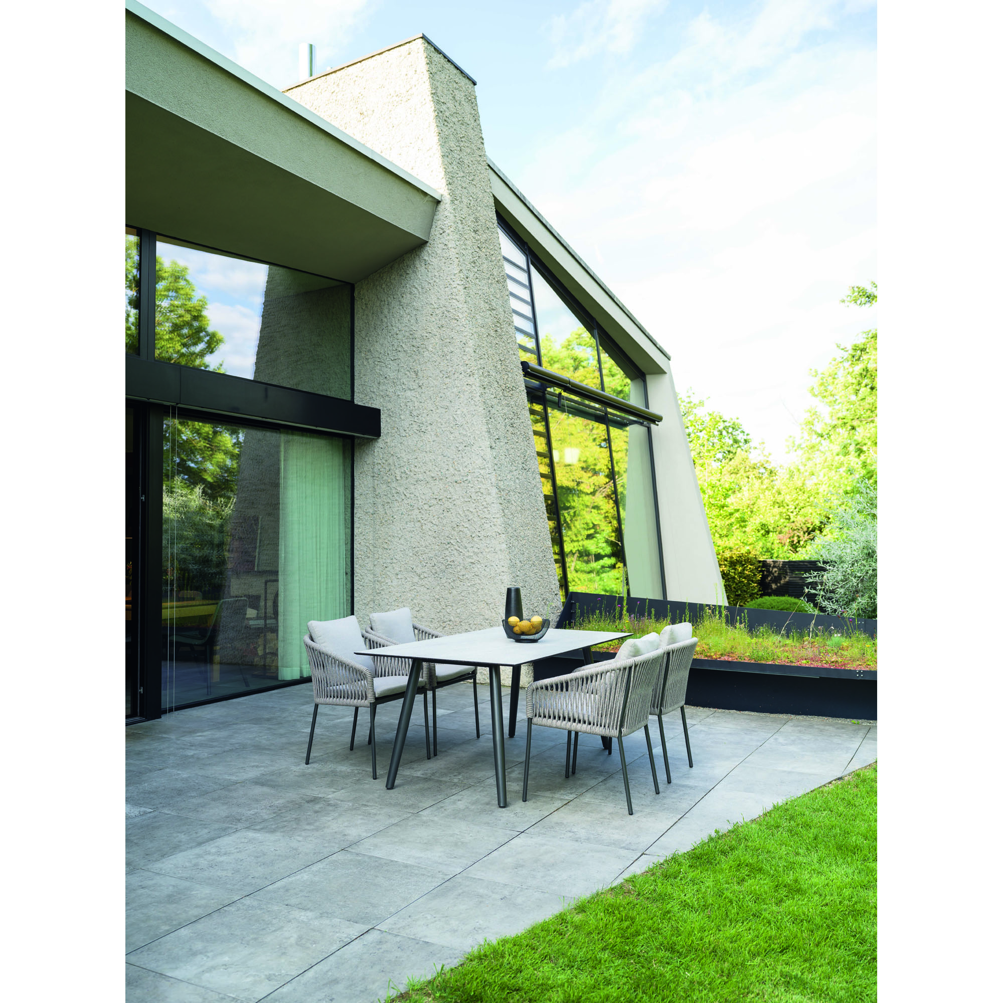 Tisch mit vier Stühlen auf einer gepflasterten Terrasse vor einem modernen Wohnhaus mit Garten
