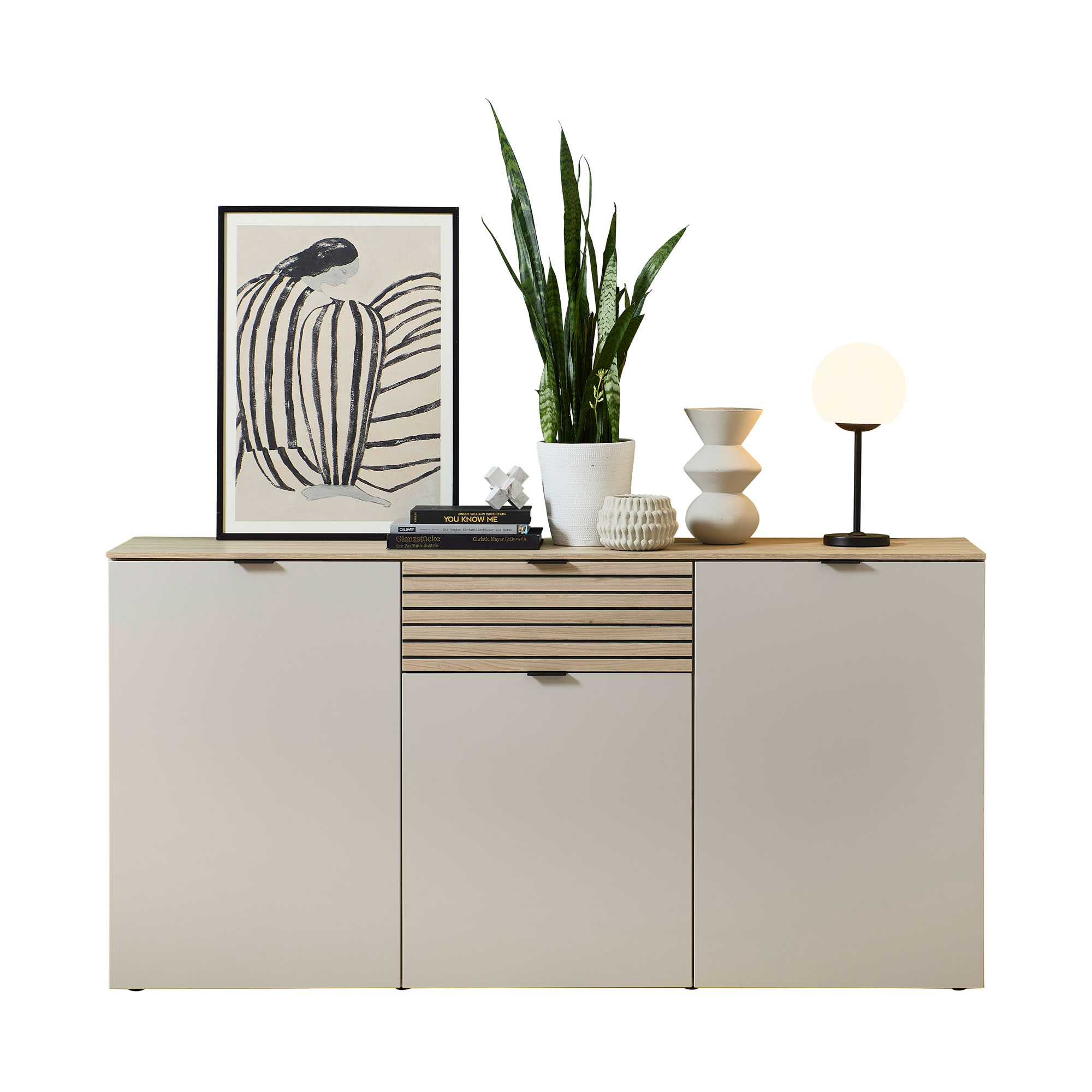 Ein Sideboard mit drei Türen und einer mittigen Schublade mit Lamellenfront. Auf der Ablagefläche stehen ein gerahmtes Bild, Bücher, eine Pflanze und Dekoobjekte.