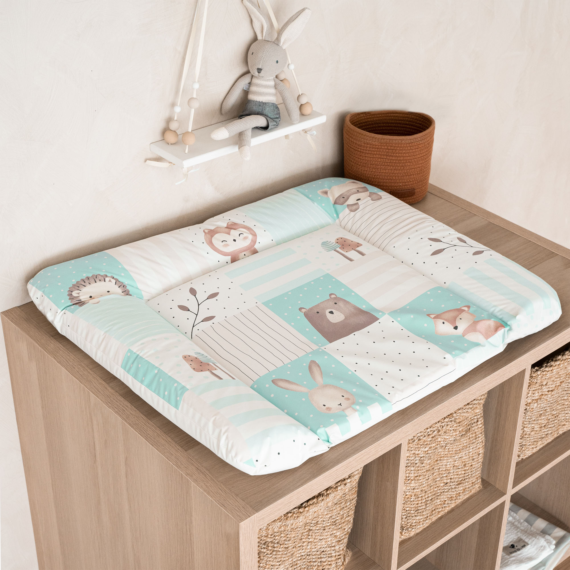 Wickelauflage mit bunten Tiermotiven in Blau-Beige auf Holzkommode mit Körben und Stoffhase im Hintergrund