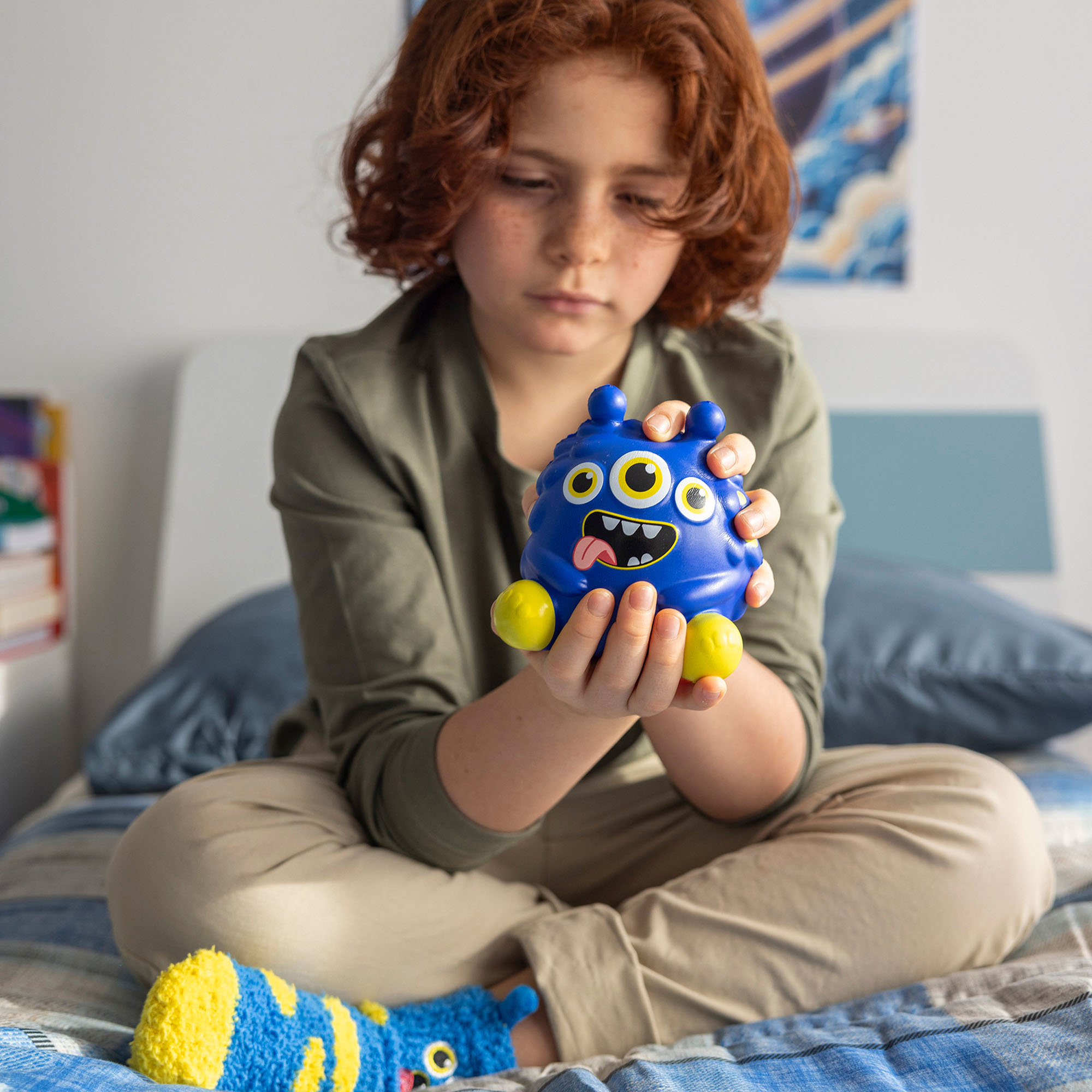 Blaue Antistress-Figur im Monster-Design, von einer sitzenden Person auf einem Bett mit beiden Händen gehalten gezeigt.