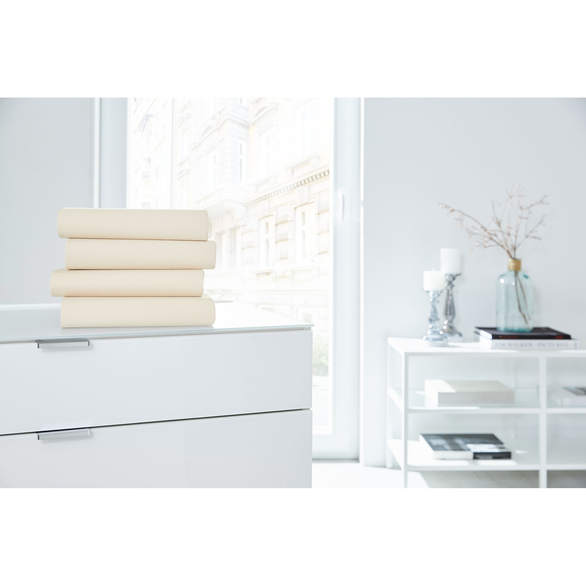 Vier gefaltete Boxspring-Spannbetttücher in Ecru auf einer weißen Kommode in einem hellen Raum; Größe 100 x 200 x 38 cm.