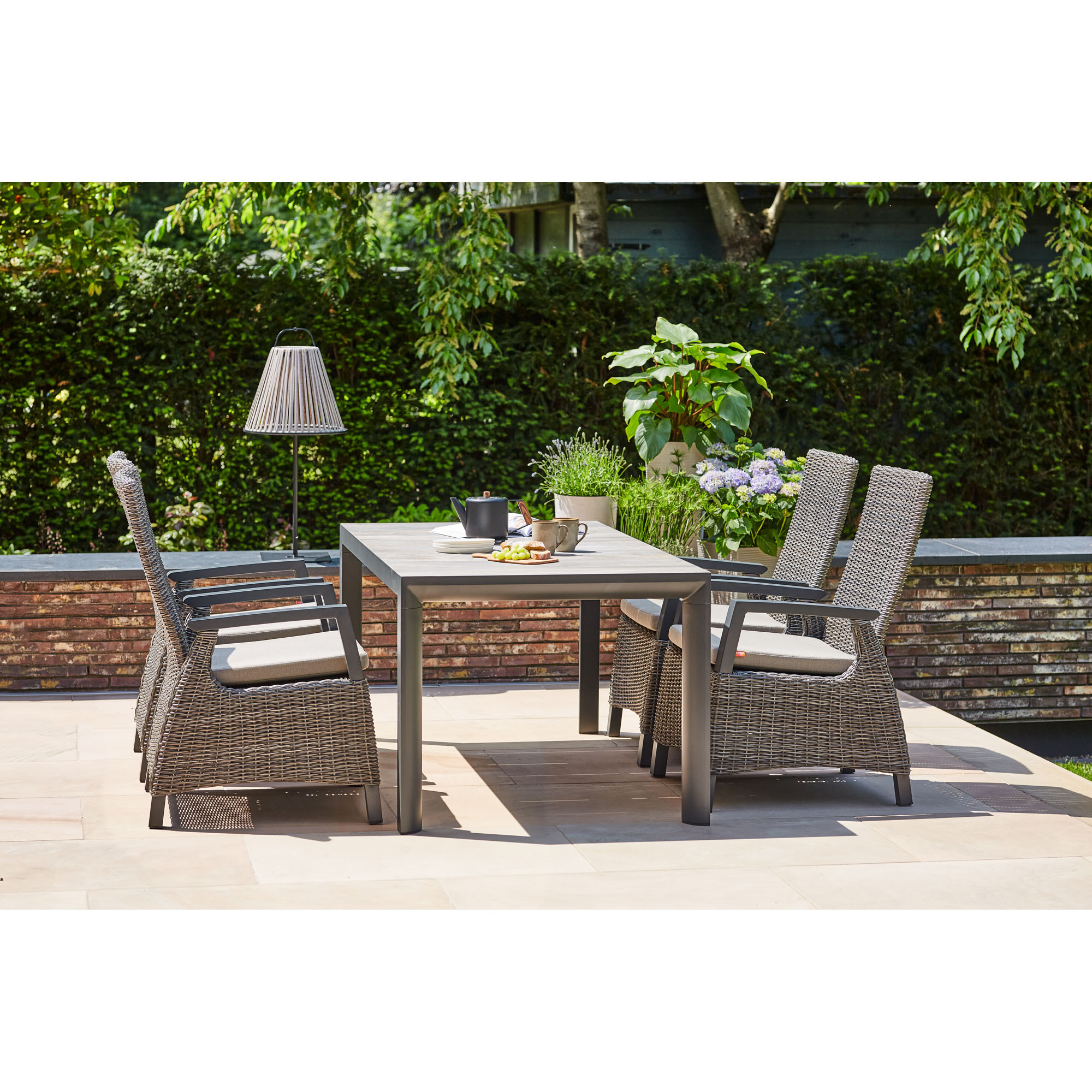 Gartentisch mit vier Gartenstühlen aus Geflecht auf einer sonnigen Terrasse, dekoriert mit Pflanzen, Getränken und Geschirr.