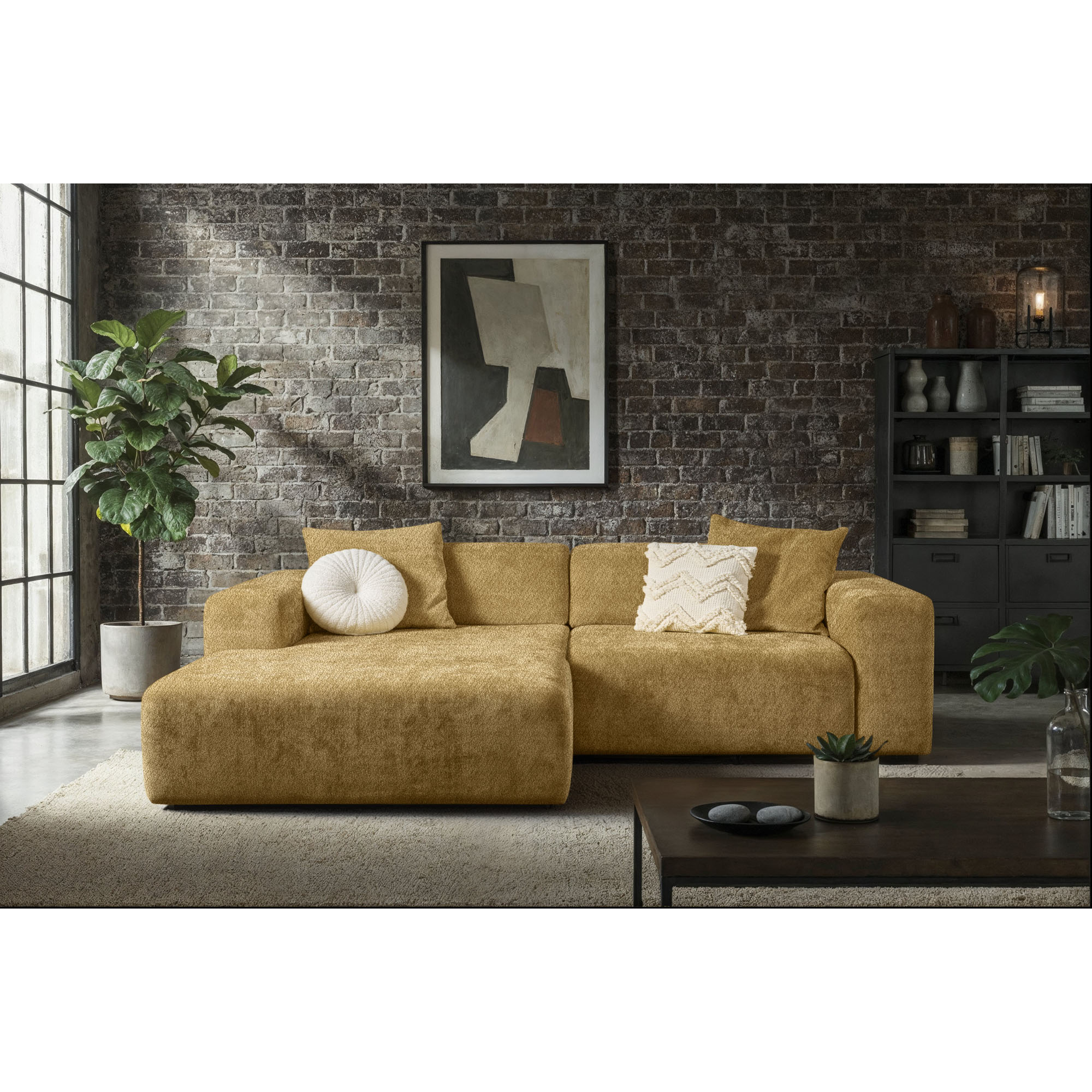 Ockerfarbene Eckcouch mit Longchair links im Bild und hellen Zierkissen vor einer dunklen Backsteinwand mit Regal und Zimmerpflanzen