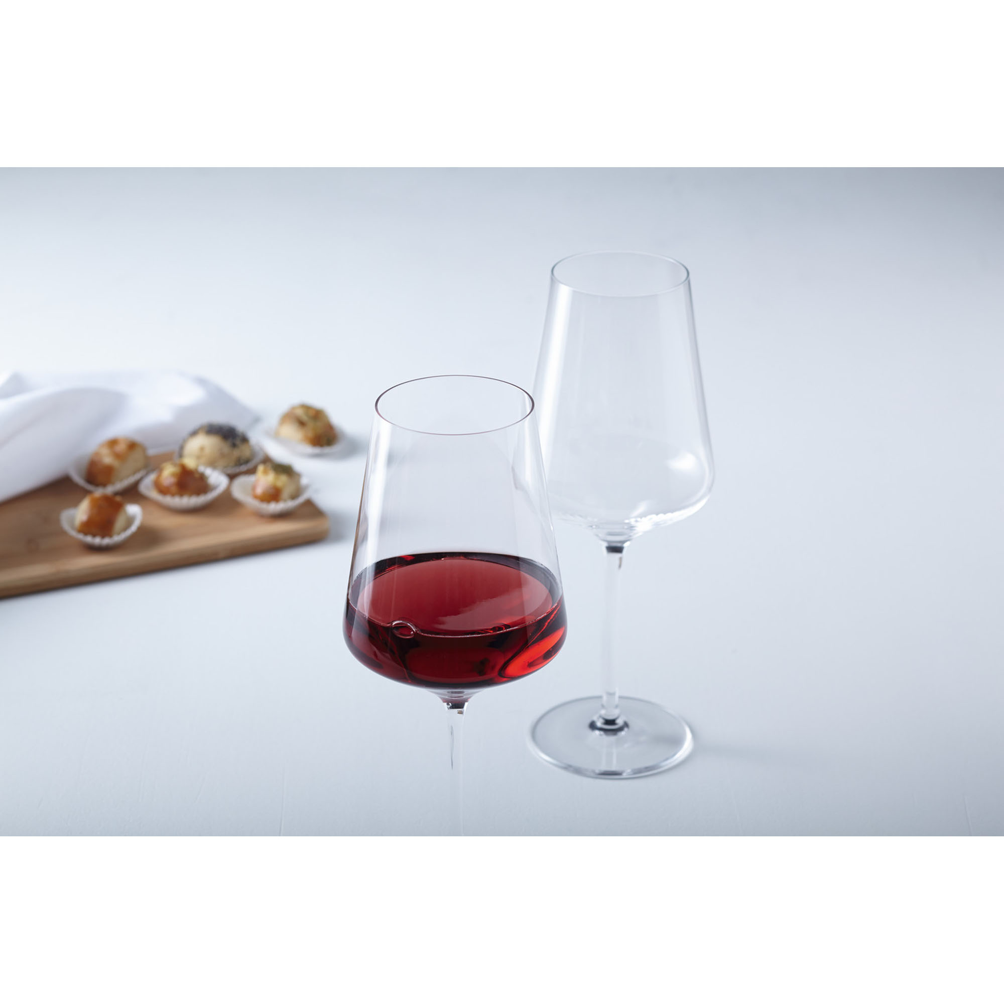 Zwei Rotweingläser eines gefüllt mit Rotwein auf hellem Tisch mit Gebäck im Hintergrund