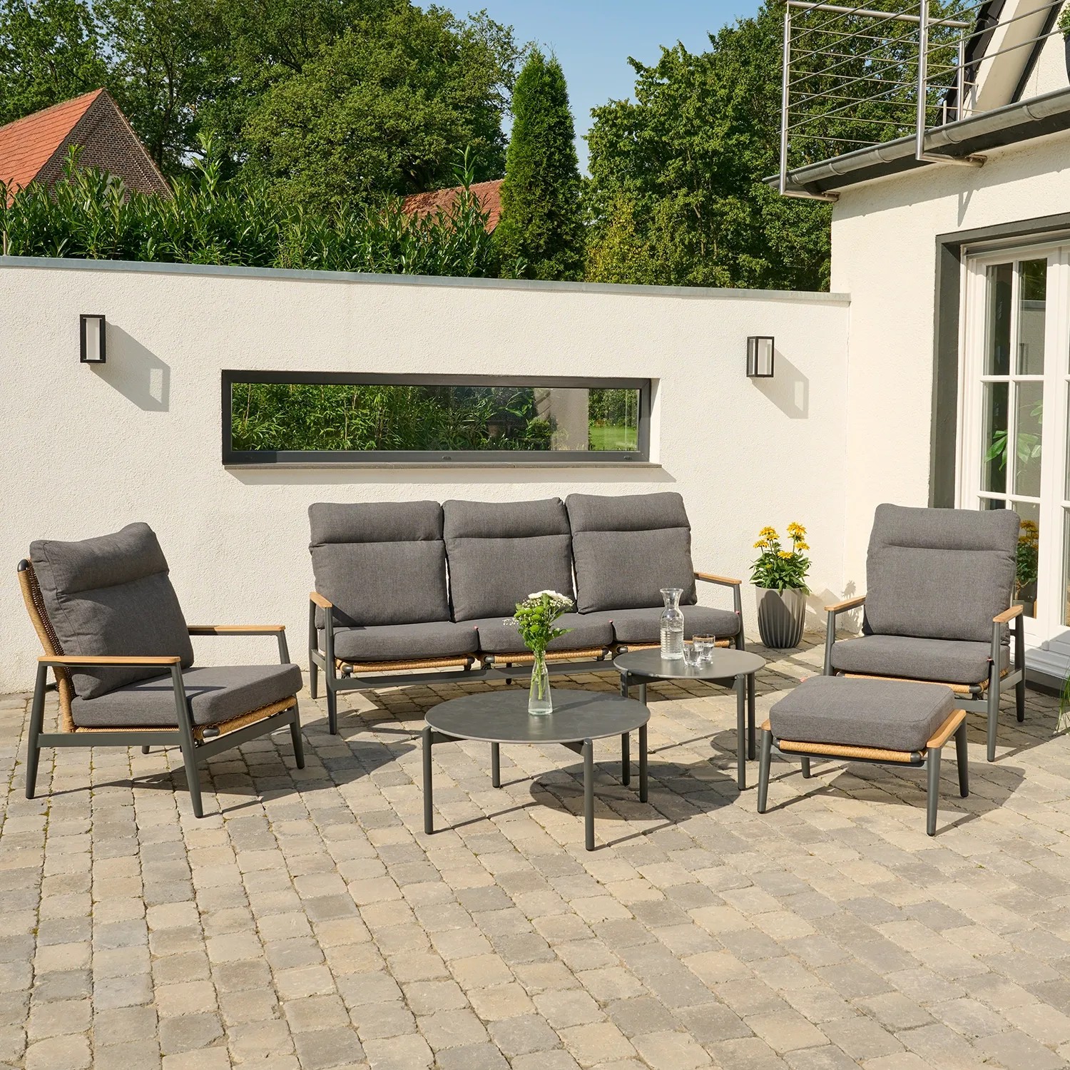 Ein Sofa mit grauen Polstern und zwei Sessel stehen auf einer gepflasterten Terrasse, ergänzt durch runde Beistelltische und Pflanzen.