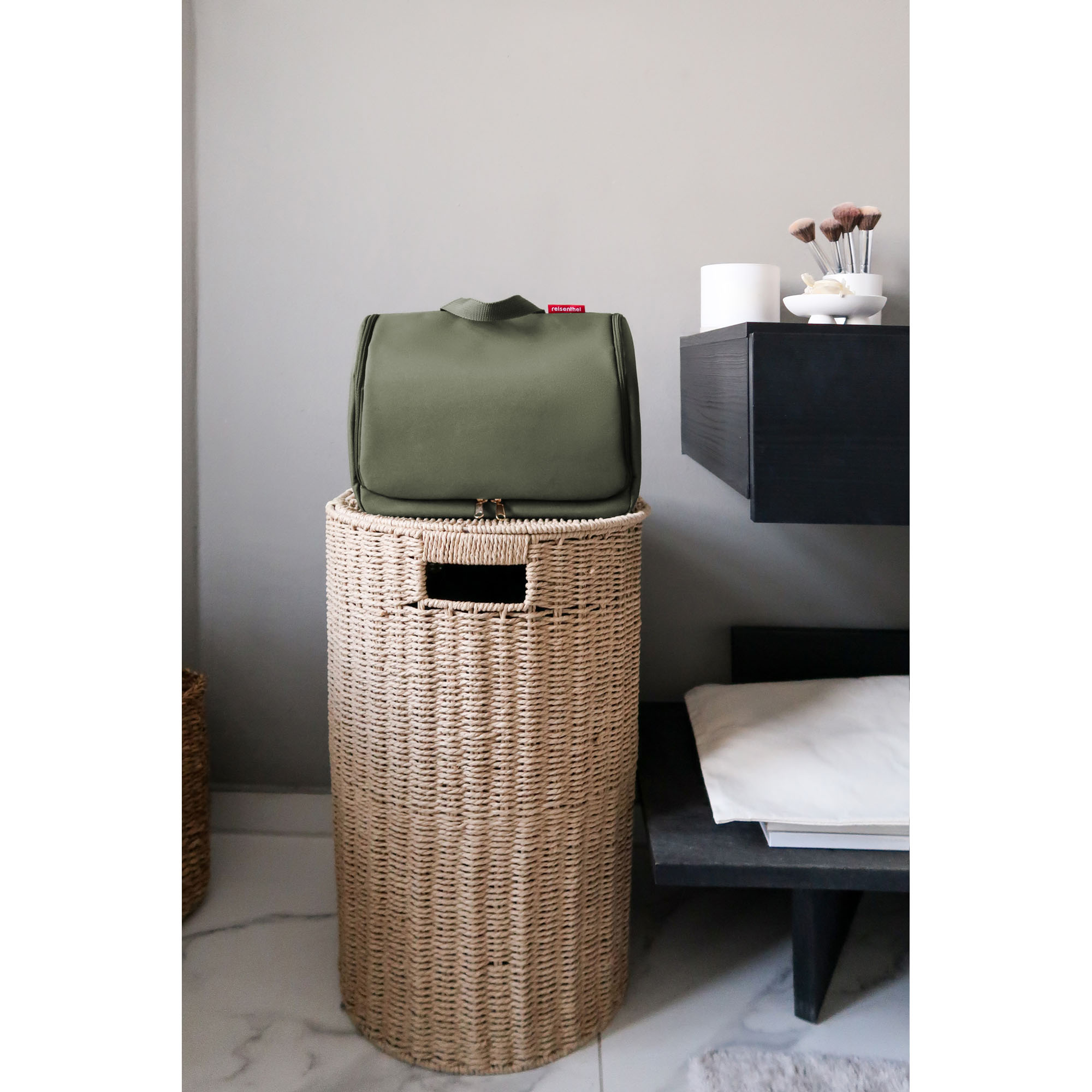 Kulturbeutel von Reisenthel, Modell Toiletbag XL, in Dunkelgrün mit goldfarbenen Metallteilen, geschlossen auf einem geflochtenen Korb in einem Wohnraum.