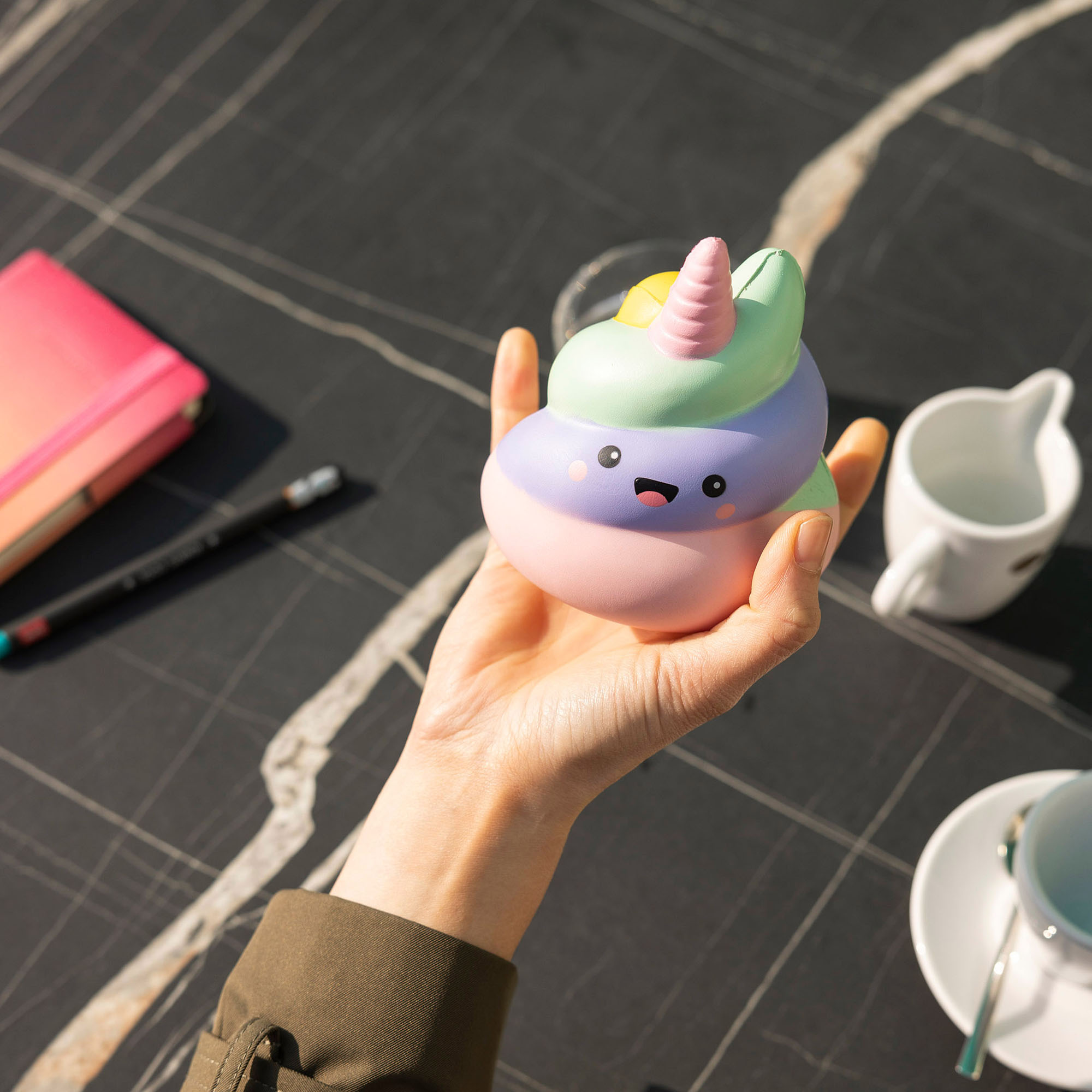Pastellfarbene Antistress-Figur in Häufchenform mit Einhornhorn, in einer Hand über einem Cafétisch mit Tassen gezeigt.