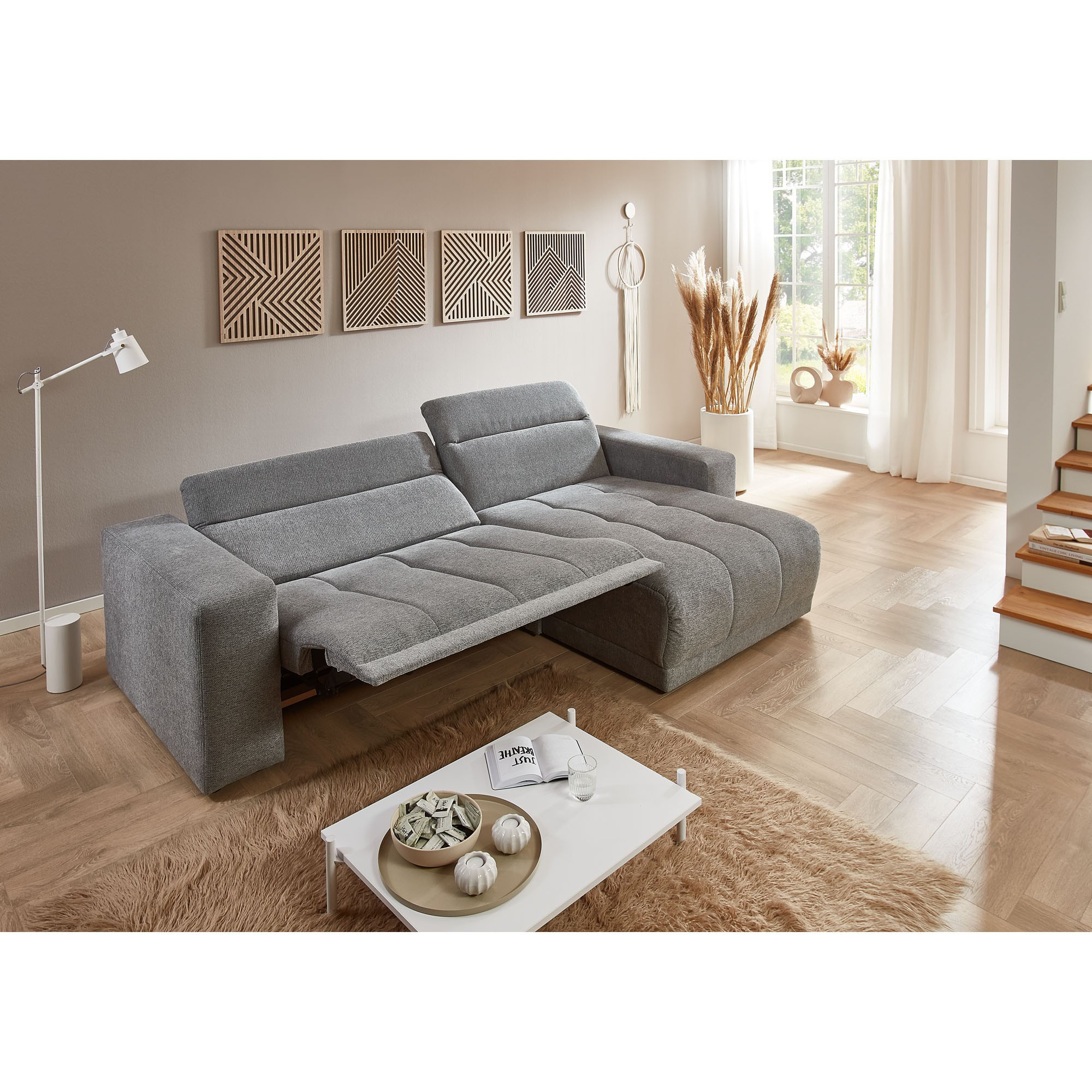 Ecksofa mit Ottomane in grauem Stoffbezug mit ausgeklappter Liegefläche und verstellbaren Rückenlehnen im Wohnraum