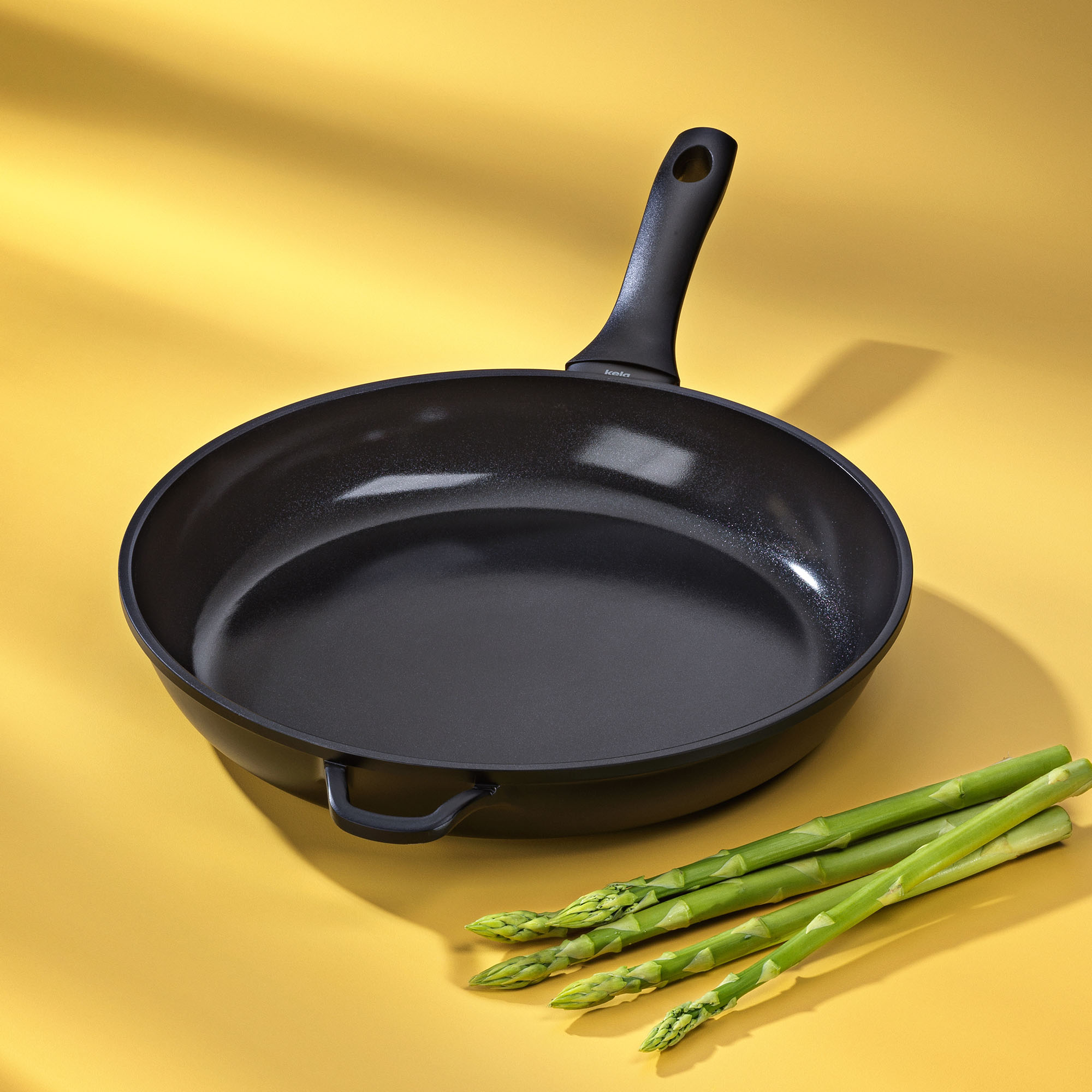 Leere schwarze Bratpfanne mit zwei Griffen neben grünem Spargel auf gelbem Untergrund