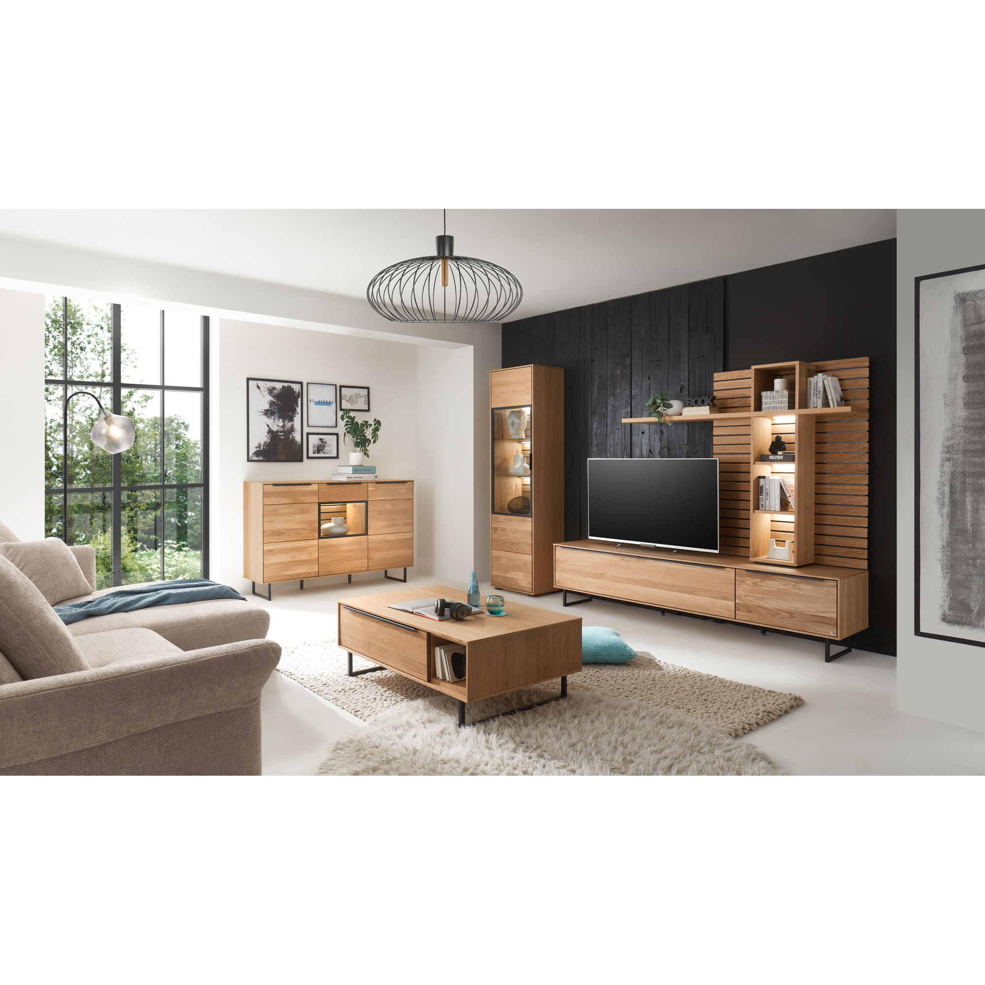 Eingerichtetes Wohnzimmer mit Möbeln der Serie Musterring Set One Jackson in Eiche teilmassiv. Die Wohnwand besteht aus Lowboard und beleuchtetem Aufsatzpaneel; zusätzlich sind Vitrine, Sideboard und Couchtisch zu sehen.