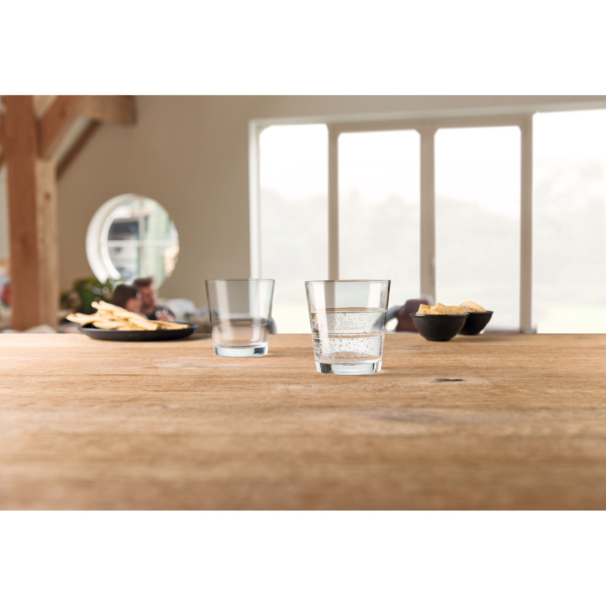 Zwei klare Trinkgläser auf Holztisch eines mit Wasser gefüllt im Hintergrund Snacks und Wohnraum
