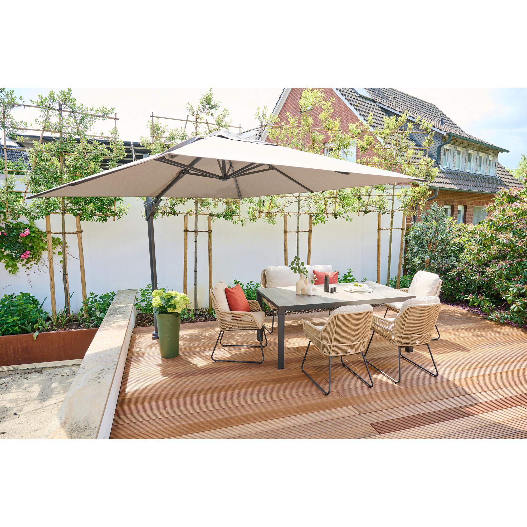 Gartentisch mit vier Stühlen auf einer Holzterrasse unter einem großen Sonnenschirm, umgeben von Pflanzen und einem Gartenzaun.
