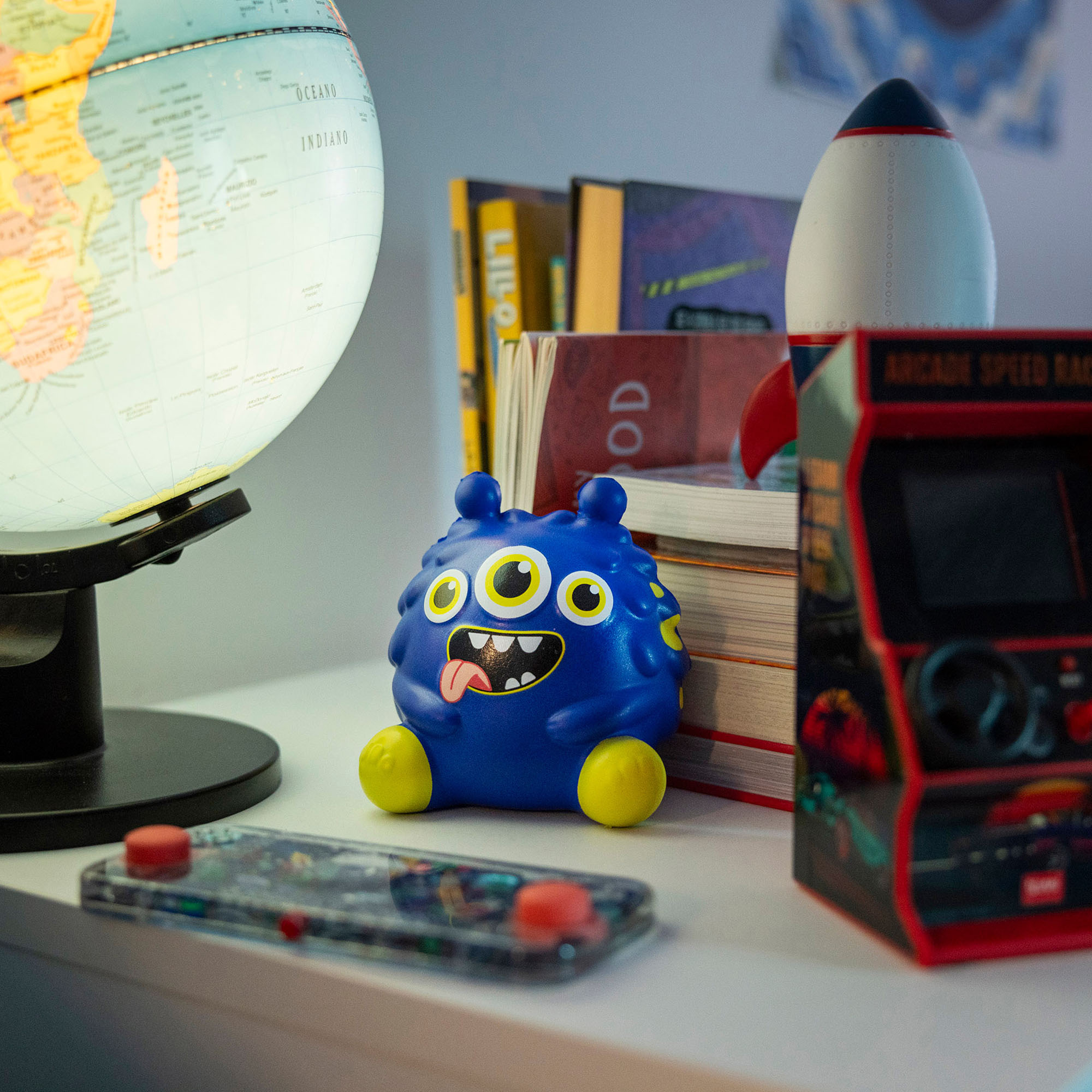 Blaue Antistress-Figur im Monster-Design, auf einem Schreibtisch neben Globus, Büchern und Mini-Arcade dargestellt.