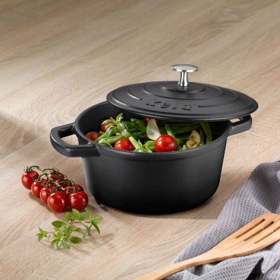 Schwarzer Bräter mit teilweise geöffnetem Deckel und verschiedenem Gemüse neben Tomaten Kräutern und Küchenutensilien