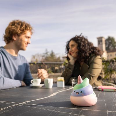 Pastellfarbene Antistress-Figur in Häufchenform mit Einhornhorn, auf einem dunklen Tisch vor zwei sitzenden Personen dargestellt.