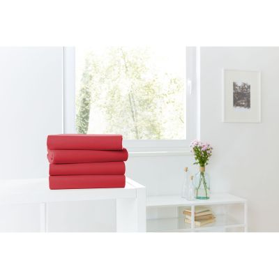 Vier gefaltete Spannbetttücher in Rot auf einer weißen Kommode vor einem hellen Fenster; Größe 100 x 200 x 30 cm.