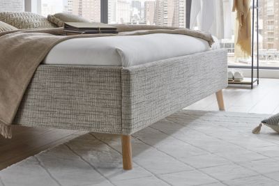 Nahaufnahme eines hellen Polsterbetts mit Holzfüßen, strukturierter Stoffbezug, weißer Bettdecke und brauner Tagesdecke in modernem Schlafzimmer