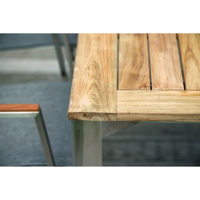 Nahaufnahme einer Tischkante mit Holzplatte in natürlicher Maserung, die auf einem silberfarbenen Metallgestell liegt. Ein Stuhl mit Holzarmlehne ist daneben zu sehen.