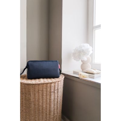Dunkelblaue Reisenthel Kosmetiktasche auf einem geflochtenen Korb neben Fenster und Dekoration