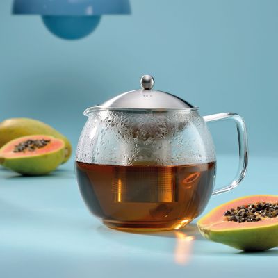Transparente Teekanne mit bernsteinfarbenem Tee zwischen zwei aufgeschnittenen Papayas auf blauem Untergrund