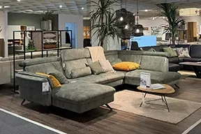 Graugrüne Wohnlandschaft mit farblich abgestimmten und gelben Kissen in einer modernen, wohnlichen Etage im Möbelhaus Möbel Gruber klein