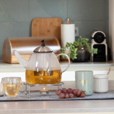 Transparente Teekanne mit Tee auf einem Tisch, daneben Tassen, Glas und Küchenutensilien