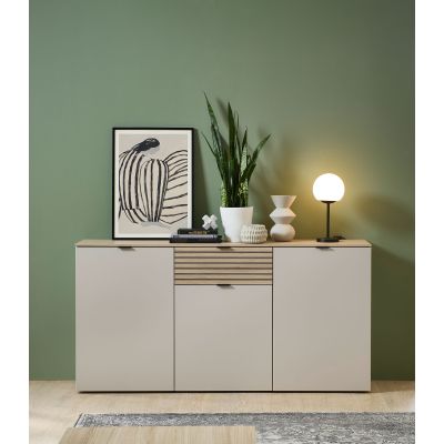 Ein Sideboard steht vor einer grünen Wand. Darauf befinden sich ein Bild, eine Pflanze, Bücher und eine Tischlampe. Der Boden ist hell und im Vordergrund ist ein Teppichausschnitt erkennbar.