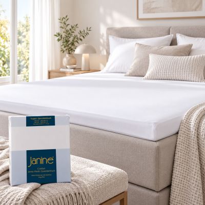 Weißes Janine Topper-Spannbetttuch 5001 glatt auf einem Topper bezogen; die Produktverpackung steht auf einer Bank vor dem Bett.