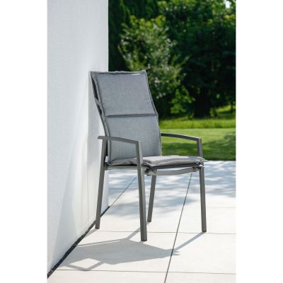 Ein grauer Gartenstuhl mit Polsterauflage lehnt an einer weißen Hauswand auf einer gefliesten Terrasse. Im Hintergrund sind grüne Bäume und Rasen im Sonnenlicht zu sehen.