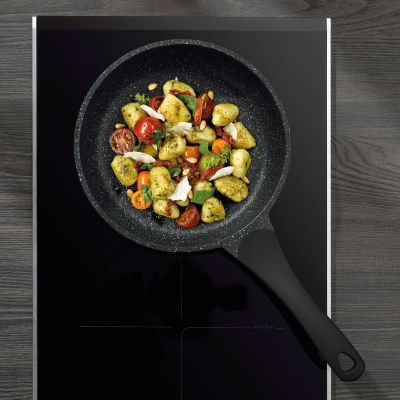 Schwarze Bratpfanne mit gewürfeltem Gemüse auf einem dunklen Kochfeld aus der Draufsicht