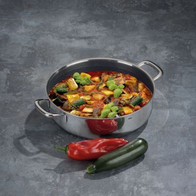 Dunkel beschichtete Servierpfanne mit buntem Gemüse auf einer grauen Arbeitsfläche neben Paprika und Chilischoten