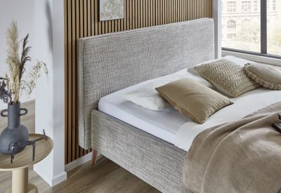 Nahaufnahme eines hellen Polsterbetts mit strukturiertem Stoffbezug, mehreren Kissen und brauner Tagesdecke vor Holzpaneelwand