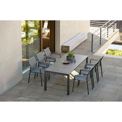 Mehrere dunkelgraue Stühle und ein rechteckiger Tisch stehen auf einer Steinterrasse neben einem modernen Haus mit Glasfront, bei Tageslicht aufgenommen.