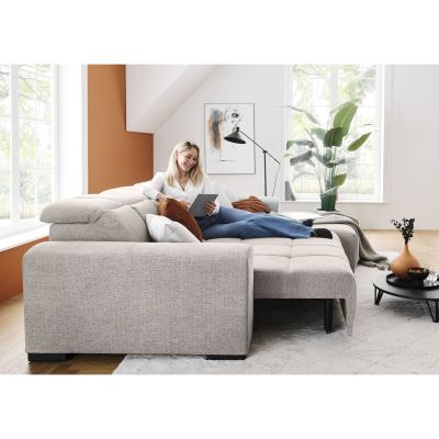 Frau liegt mit einem Tablet auf einem silbergrauen Schlafsofa mit ausgezogener Liegefläche, erhöhten Kopfstützen und mehreren Kissen im Wohnzimmer
