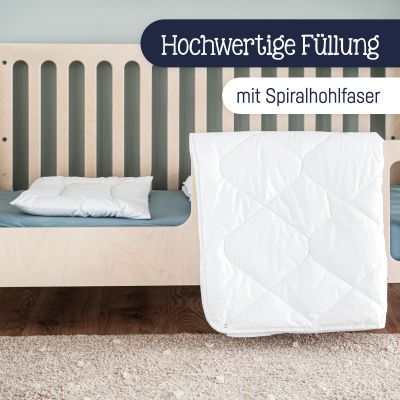 Weiße Steppdecke über Bettkante gehängt und weißes Kissen im Kinderbett mit Holzrahmen platziert