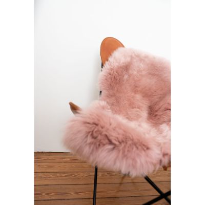 Rosafarbenes Lammfell liegt über Sitz und Lehne eines orangebraunen Sessels mit schwarzem Metallgestell auf Holzdielen.