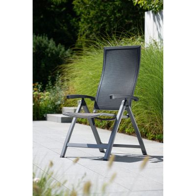 Ein dunkelgrauer Gartenstuhl mit hoher Rückenlehne steht auf einer hellen Terrasse, umgeben von hohen grünen Gräsern und Pflanzen im Hintergrund.