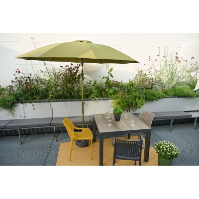 Ein quadratischer Tisch mit Stühlen steht auf einer Terrasse, darüber ein grüner Sonnenschirm. Im Hintergrund sind Pflanzen und Sitzbänke.