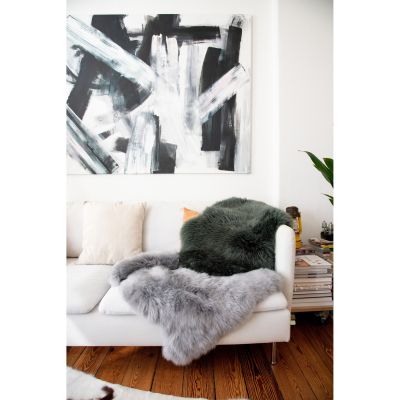 Dunkelgrünes Fell liegt an der Rückenlehne eines hellen Sofas; ein graues Fell hängt über die Sitzkante, darüber ein großes Wandbild.