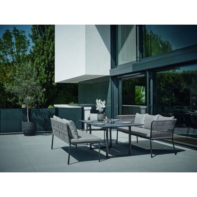 Eine Terrassenszene mit zwei Gartenbänken, einem Tisch und einem Baum im Topf, vor einem modernen Gebäude mit großen Glasflächen.