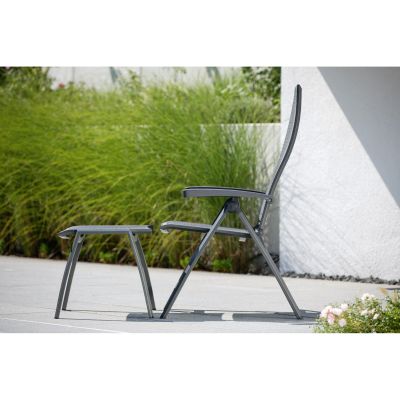 Ein seitlich fotografierter Gartenstuhl mit Hocker aus grauem Metall und Textilbespannung. Im Hintergrund sind hohe Gräser und ein heller Hausputz zu sehen.
